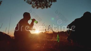 两名游客正坐在篝火晚会上，在大自然中露营，剪影，阳光日落。 两个男人剪影